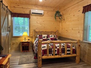 A wooden cabin interior with a bed, a nightstand, a lamp, and a window with blinds. - Woodland Cabin Retreat Near Amish Country (Warsaw)