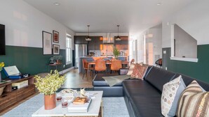 Una sala de estar moderna con un sofá de color oscuro, una mesa de centro con libros y un jarrón de flores, un comedor con una cocina al fondo, y un ventilador de techo.