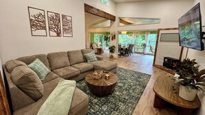 Ein geräumiges Wohnzimmer mit einem braunen Ecksofa, einem Holztisch und einem Teppich mit grünem Muster. Durch eine Glastür ist ein Essbereich mit Tisch und Stühlen zu sehen.