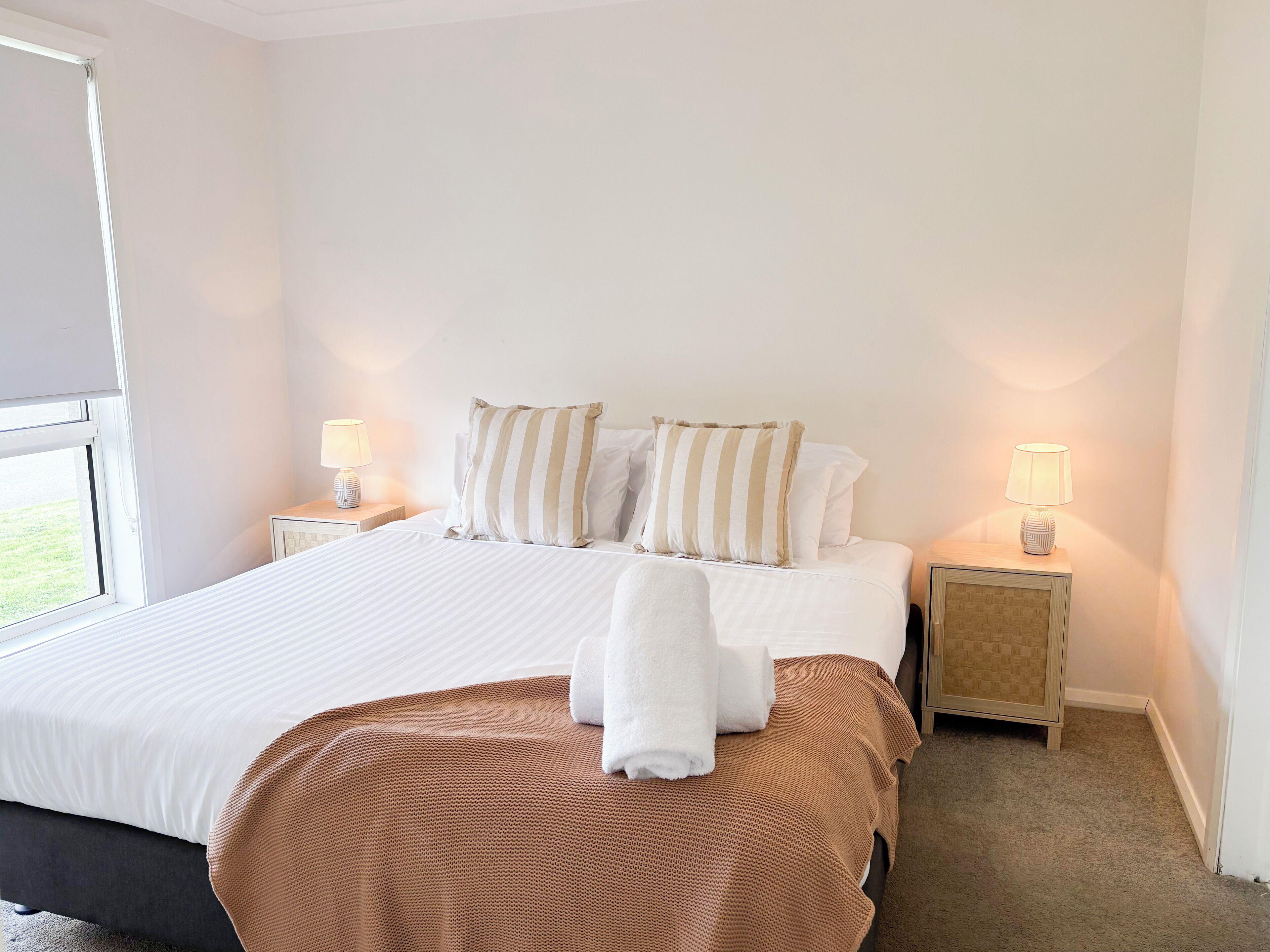 Habitación de hotel con una cama grande, dos mesitas de noche con lámparas, un ventanal y un banco marrón con textura.