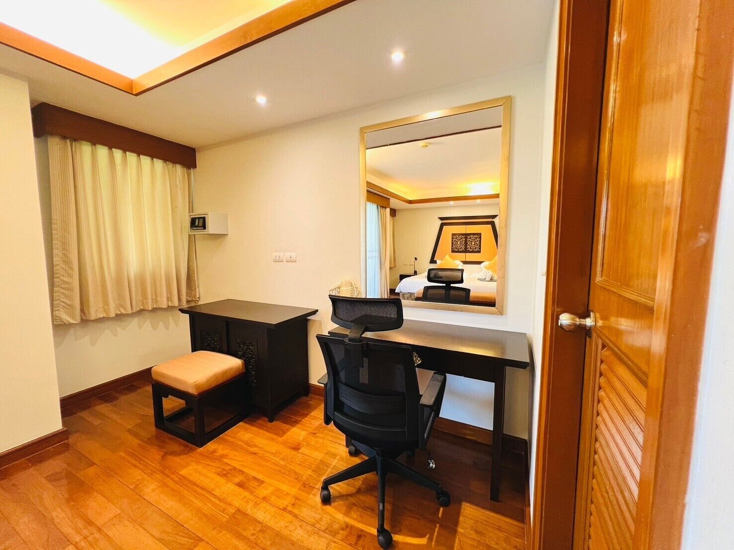 Habitación de hotel con un escritorio de madera, una silla, un pequeño banco, un espejo y una puerta.