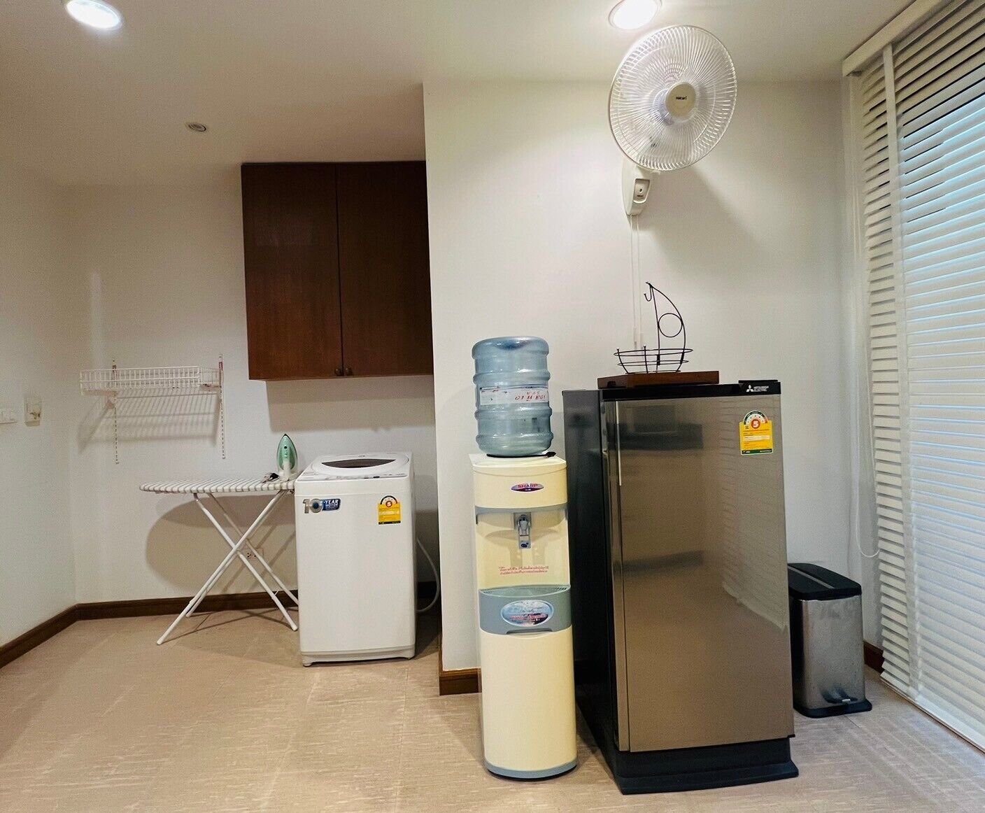 A compact kitchen with a refrigerator, a water dispenser, a fan, and a drying rack.