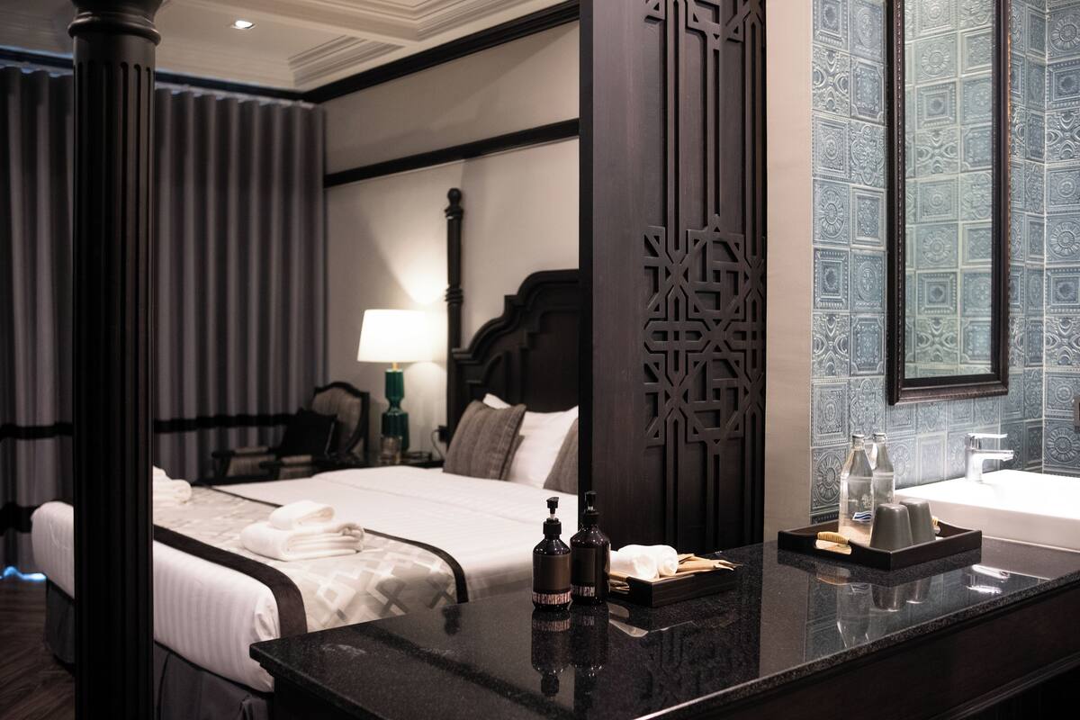 A hotel room with a neatly made bed, a bedside table with toiletries, a large mirror, and a bathroom with a sink and towel rack.