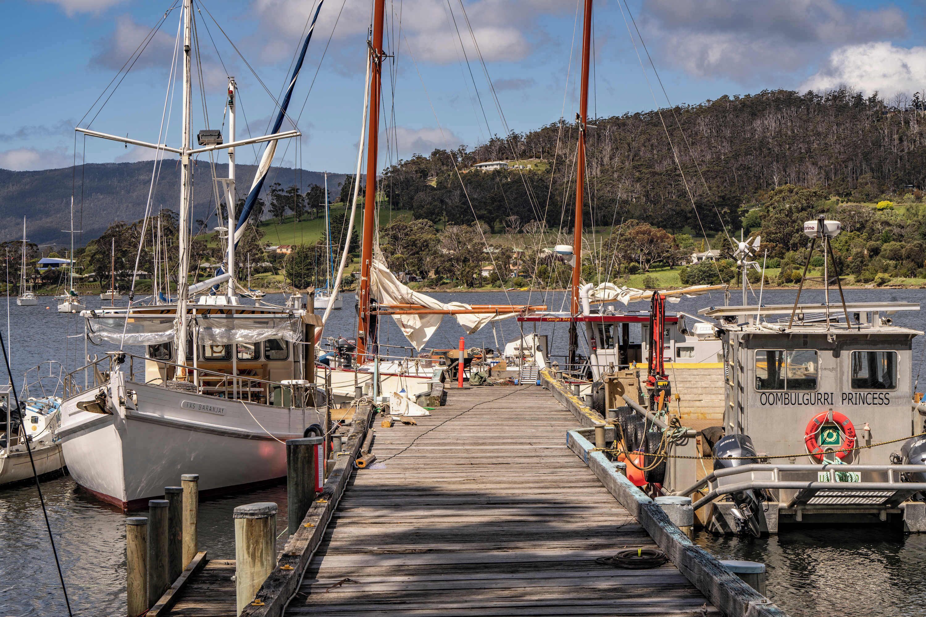 A wooden dock with boats moored at a marina, surrounded by hills and a clear sky.