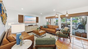 A spacious living room with a leather sofa, a green armchair, and a coffee table. There is a TV on the wall and a large window providing a view of the outdoors.