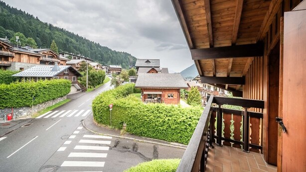 A traditional wooden building with a balcony, a crosswalk, and a road leading to a small house surrounded by greenery.