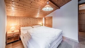 A bedroom with a wooden ceiling, a bed with white bedding, a nightstand with a lamp, and a door to another room.