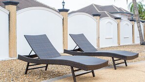 Two wicker lounge chairs with metal frames, positioned on a gravel surface, adjacent to a white wall with decorative arches and a tiled roof.