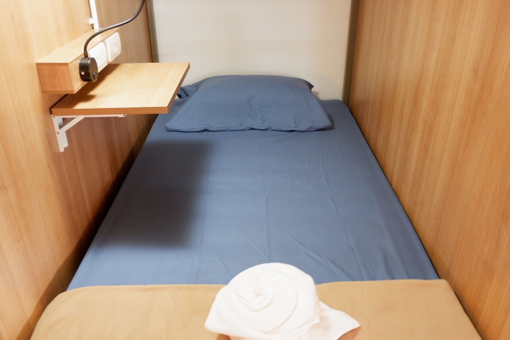 A small, single-bed room with a wooden bunk bed structure, a pull-out desk, and a wall-mounted power outlet.