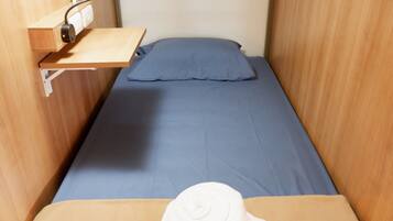 A small, single-bed room with a wooden bunk bed structure, a pull-out desk, and a wall-mounted power outlet.