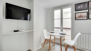 A modern dining area with a round table, white chairs, a flat-screen TV, and framed pictures on the wall. - Intimate Apartment - 2P - Le Marais (Paris)