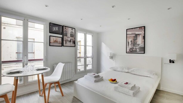 A modern hotel room with a large bed, a dining area with a table and chairs, and a view of the street through large windows. - Intimate Apartment - 2P - Le Marais (Paris)