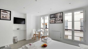 A modern hotel room with a bed, a dining area with a table and chairs, a TV mounted on the wall, and large windows offering a view of the city. - Intimate Apartment - 2P - Le Marais (Paris)