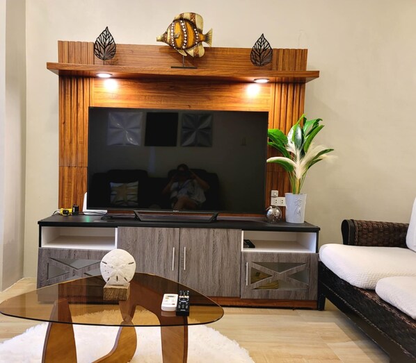Un salon moderne avec un grand téléviseur à écran plat fixé sur un mur en bois, une table basse en verre, un canapé blanc et une plante en pot.