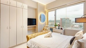 Un hotel moderno con una cama grande, un televisor, una lámpara de pared redonda y vistas de la ciudad a través de un ventanal amplio.
