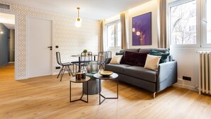 A modern living room with a grey sofa, a dining table with chairs, and a large window with curtains.