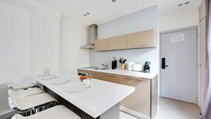 A modern kitchen with white countertops, beige cabinets, and a stainless steel sink. There is a dining area with a white table and chairs, and a door with a sign on it. - Cosy Apartment - 1br/4p - Gare Saint-lazare (Paris)