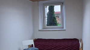 Une pièce avec un plafond en pente, une fenêtre donnant sur un arbre et un immeuble, un lit recouvert d’une couverture violette, une chaise avec un coussin bleu et une table avec un plateau en verre.