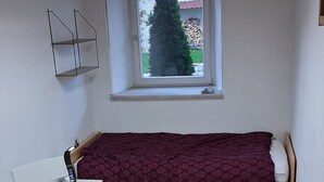 Une pièce de taille modeste comprenant un lit, une petite table, une chaise, une fenêtre donnant sur des arbres et un luminaire au plafond.
