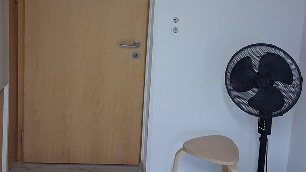 Une pièce avec une porte en bois, un petit tabouret, un ventilateur sur pied et un mur blanc équipé de prises électriques.