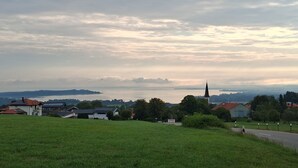 Un paysage rural avec des maisons, une église et une vue sur un lac et des collines lointaines.