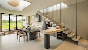 A modern kitchen with a central island, a staircase, and large windows offering a view of greenery. - Villa Mesra Stunning Paddy Fields Views - Canggu Berawa (Bali)