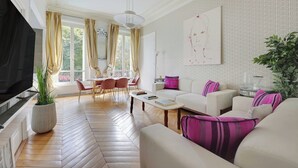 A spacious living room with a large TV, a dining area with chairs, a sofa with cushions, and a large window with curtains. - Charming 2br/4p - Georges V / Champs-elysees (Paris)