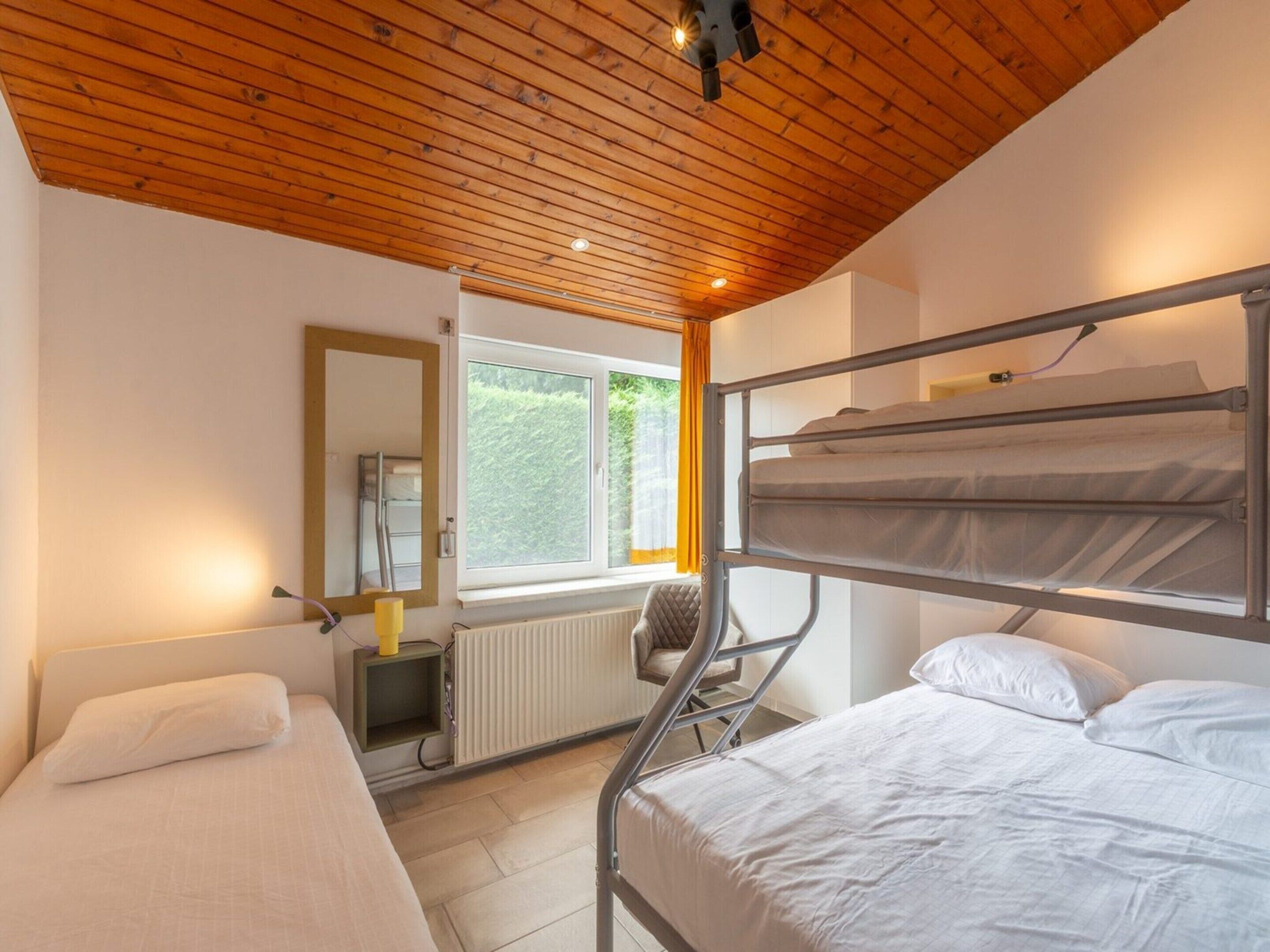 Een slaapkamer met een stapelbed, een enkel bed, een radiator, een raam met uitzicht op groen, en een houten plafond.