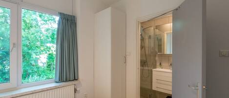 A bedroom with a bed, a window with curtains, a radiator, and a door leading to a bathroom with a shower and a mirror.
