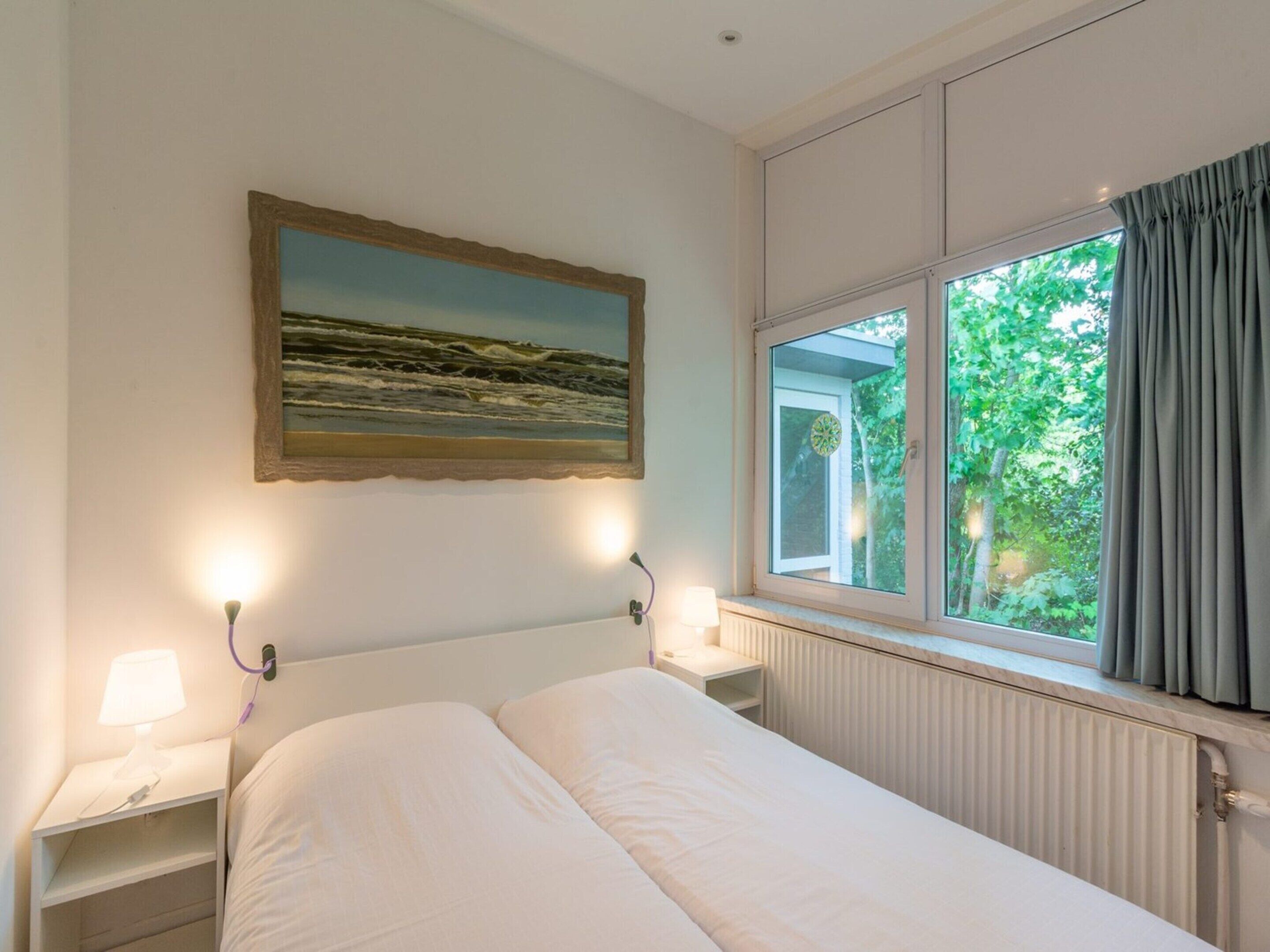 A bedroom with a bed, a nightstand with a lamp, a window with curtains, and a painting on the wall.