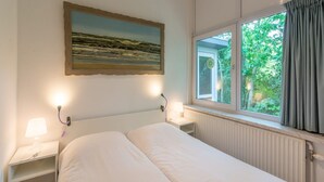A bedroom with a bed, a nightstand with a lamp, a window with curtains, and a painting on the wall. - Holiday Home in Sint Maartenszee (Sint Maartensvlotbrug)