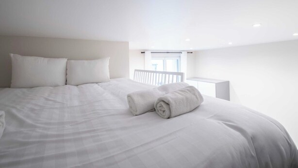 A large bed with white linens, a white headboard, and two rolled towels on the bed.