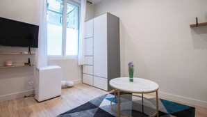 A modern living room with a flat-screen TV, a white wardrobe, a round table with a vase, and a patterned rug.