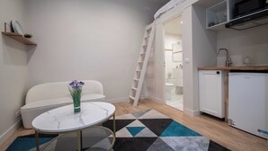A compact living space with a sofa, a small round table, a vase with flowers, a geometric-patterned rug, a white refrigerator, a white dishwasher, and a kitchen area with a microwave and a sink.
