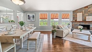 A spacious living room with a dining area, large windows with striped curtains, a fireplace, and a ceiling fan.