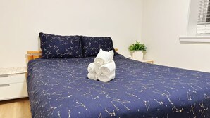 Cama bem arrumada com colcha azul escura estampada com um padrão de constelações, um travesseiro branco e uma toalha branca enrolada.
