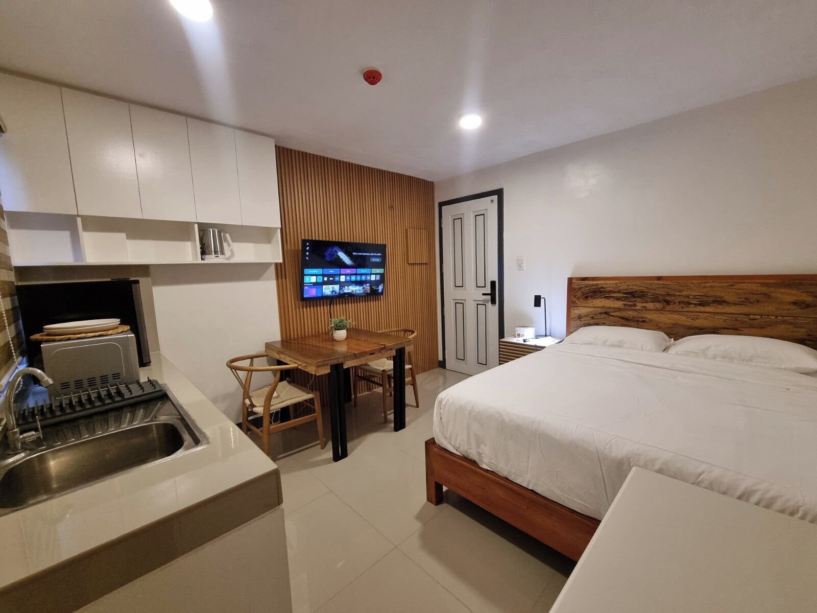 A compact hotel room with a kitchenette, a dining area, a bed, and a TV mounted on the wall.