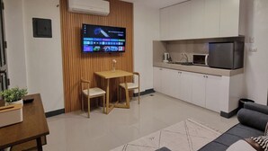 A modern living room with a sofa, a dining table with chairs, a TV mounted on a wooden wall, and a kitchen area with cabinets and appliances. - Flat 35  (Las Piñas)