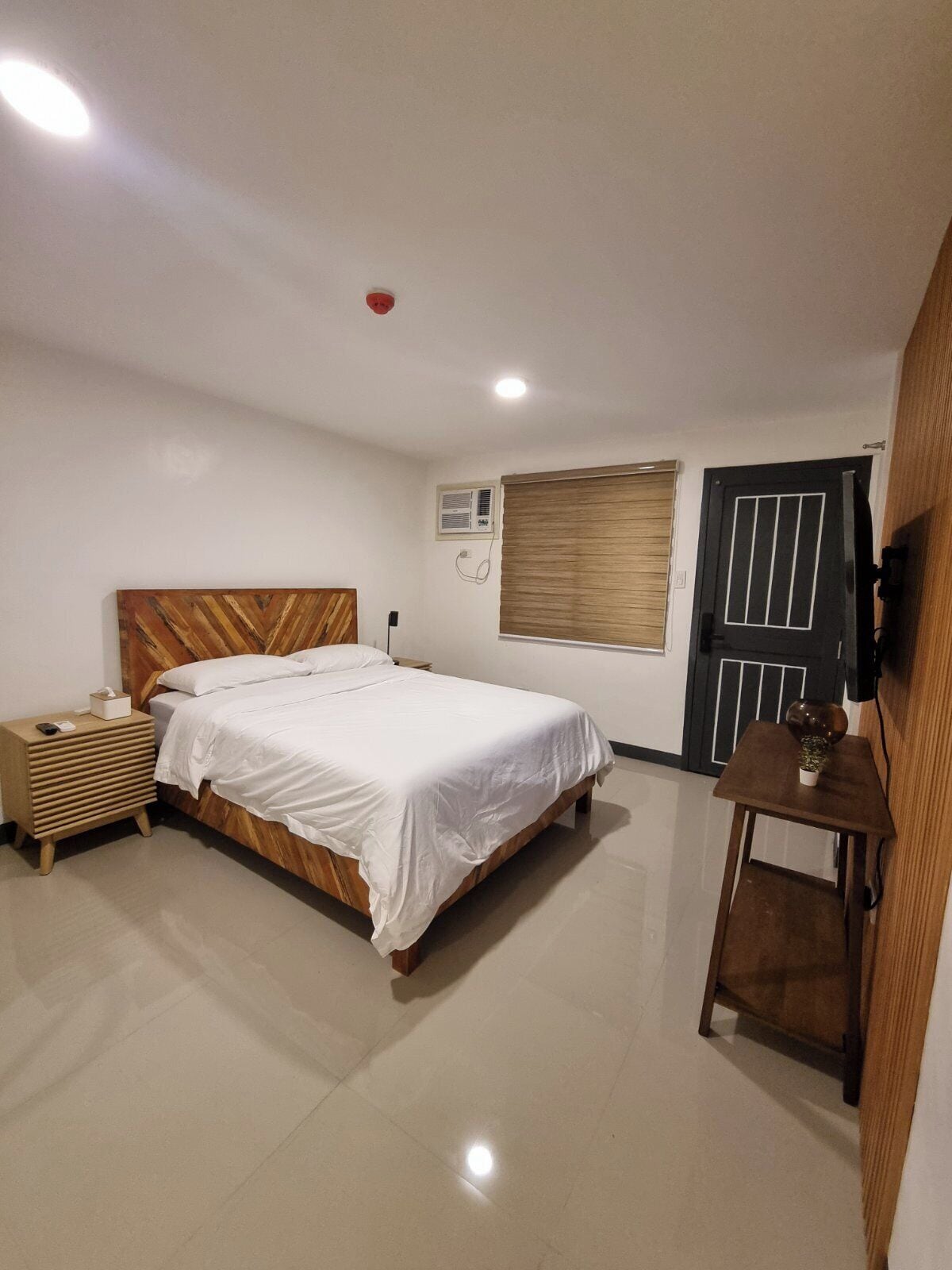 A bedroom with a wooden headboard bed, a bedside table with a lamp, a small table with a plant, and a door with a window.