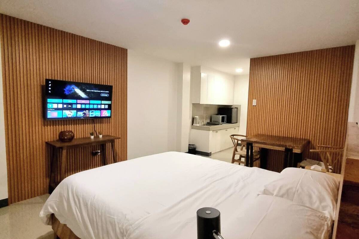 Ein Hotelzimmer mit einem großen Bett, einem an der Wand montierten Fernseher, einer Wand mit Holzpaneelen und einer Küchenzeile im Hintergrund.