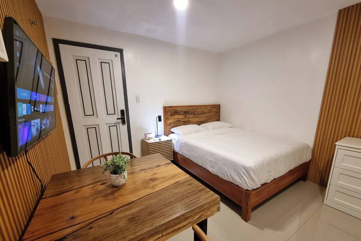 Ein Schlafzimmer mit einem hölzernen Bett, einem Nachttisch, einem Schreibtisch aus Holz und einem Topf mit einer Pflanze. An der Wand ist ein Fernseher angebracht.