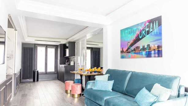 A modern living room with a teal sofa, a cityscape artwork, and a kitchen area in the background. - Modern Apartment - 1br/4p - Pereire (Paris)