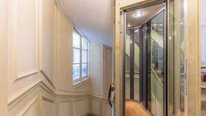 A hallway with a glass elevator door, a carpeted staircase, and a window with white paneled walls. - Modern Apartment - 1br/4p - Pereire (Paris)