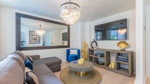 Una sala moderna con un espejo grande, una araña, un sillón azul, una mesa de centro redonda y una estantería.