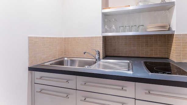 Una cocina con un lavamanos de acero inoxidable, una encimera negra y una estufa eléctrica empotrada. Hay azulejos color beige en la salpicadera y una repisa sobre el lavamanos con cristalería y platos.
