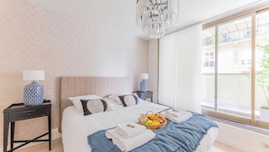 Habitación de hotel moderna con una cama grande, dos mesitas de noche con lámparas, una araña y vistas a la ciudad a través de grandes ventanales.