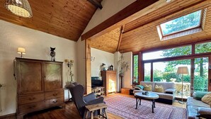 A spacious living room with wooden floors, a large wooden cabinet, a comfortable sofa, a television, and a large window offering a view of greenery outside. - An oasis of tranquility in the heart of nature (Rendeux)