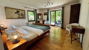 A bedroom with a wooden bed, a nightstand with a lamp, a chair, a desk, and a view of the outdoors through a glass door. - An oasis of tranquility in the heart of nature (Rendeux)