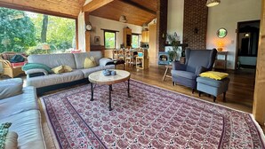A spacious living room with a large sofa, a coffee table, and a patterned rug. There is a fireplace, a dining area, and a kitchen visible in the background. - An oasis of tranquility in the heart of nature (Rendeux)