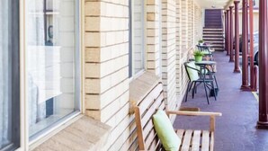 Un espace extérieur abrité, doté de bancs et de chaises en bois, d’un mur en briques et offrant une vue sur un bâtiment en arrière-plan.